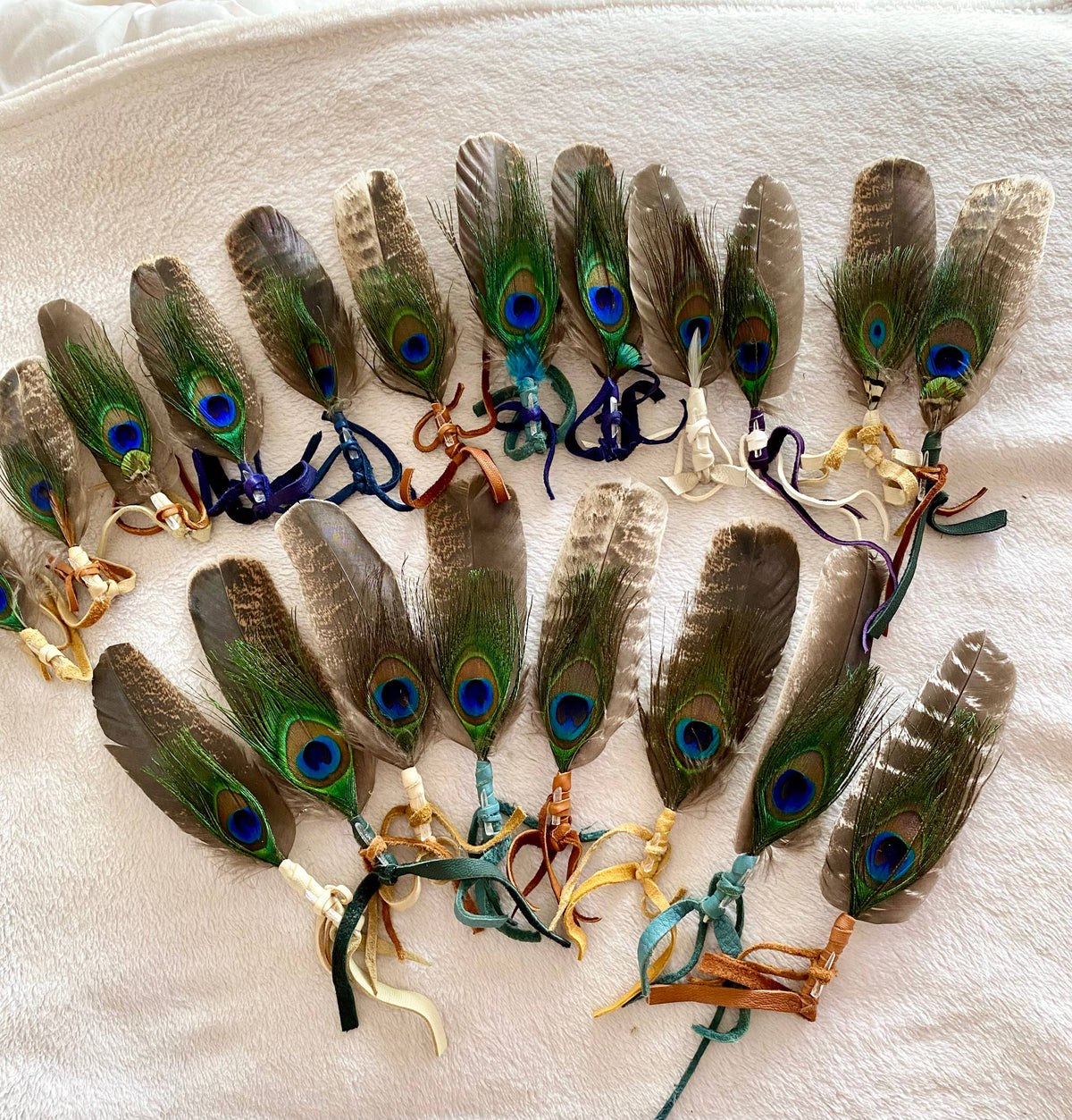 A set of twelve peacock feather wristlets or keychains arranged in a semi-circle on a textured white fabric background. Each feather features a natural green, brown, and blue eye pattern, finished with a colorful ribbon tie in shades of blue, teal, brown, and cream.