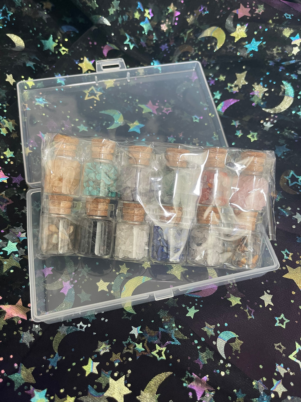 A clear plastic case containing multiple small glass vials with corks, filled with colorful powder or resin pieces, photographed on a black background printed with iridescent stars and crescent moons.
