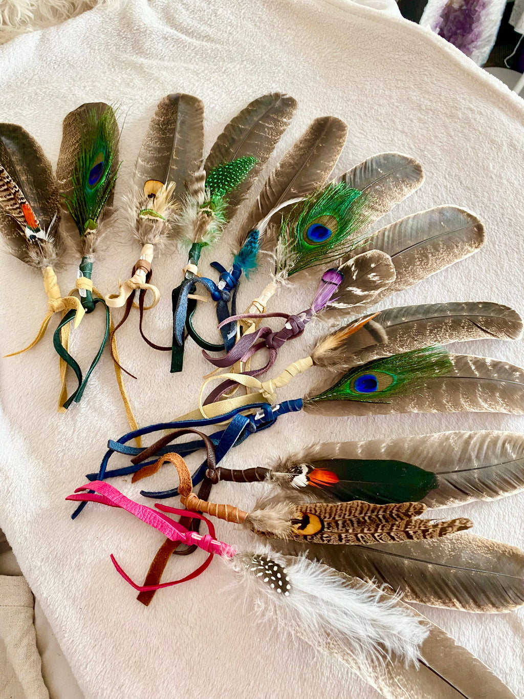 A collection of handmade feather accessories with colorful ribbons, laid out on a light beige fabric background. The set includes various brown and gray feathers, green peacock feathers with blue eyes, white feathers, and bright pink, blue, purple, and green ribbons.