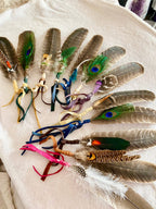 A collection of handmade feather accessories with colorful ribbons, laid out on a light beige fabric background. The set includes various brown and gray feathers, green peacock feathers with blue eyes, white feathers, and bright pink, blue, purple, and green ribbons.